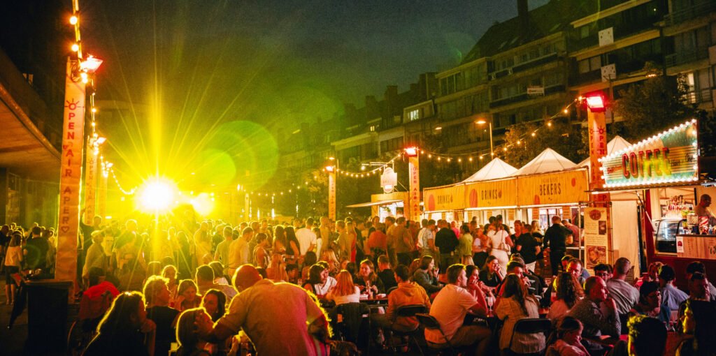 Avondbeeld van het verlichte terrein van Open Air Apero met volle tafels en gezellige sfeer.