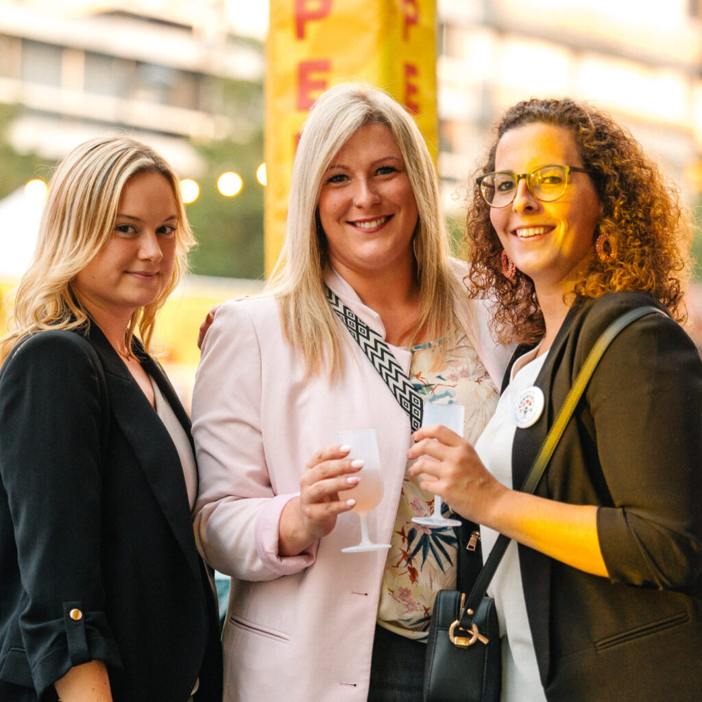 Drie feestelijk geklede vrouwen poseren met drankjes bij Open Air Apero – afterwork en uitgaan in stijl.