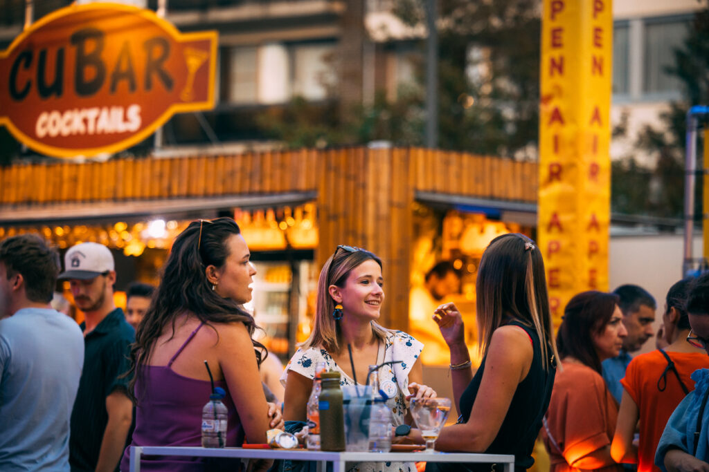 Drie vrouwen praten en lachen aan een statafel voor de Cubar cocktailbar op Open Air Apero.